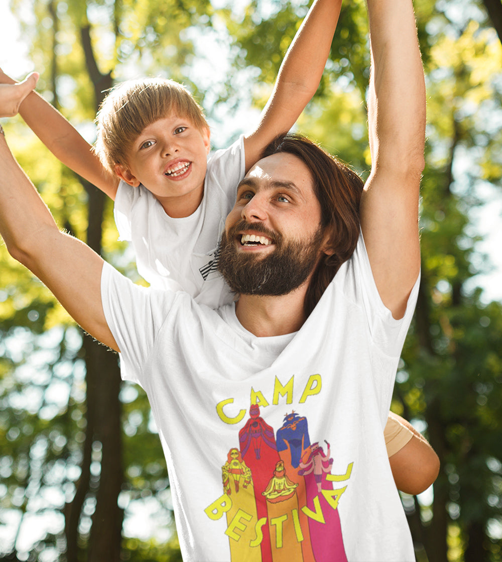 A smiling man carries a happy young boy on his shoulders outdoors. Both wear white t-shirts; the man’s shirt says “Camp Bestival” with colorful cartoon figures. Sunlight filters through green trees.