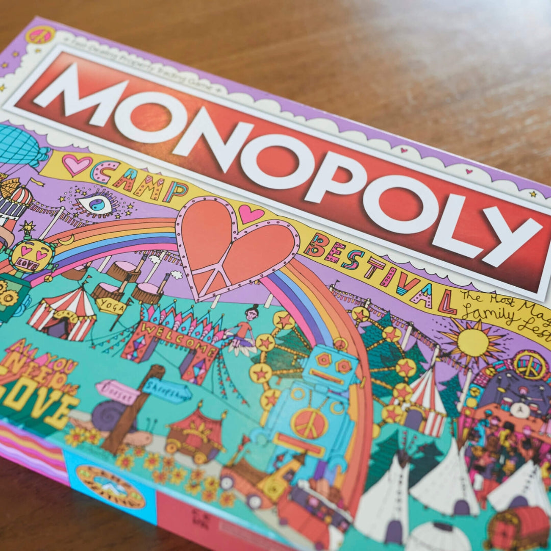The Camp Bestival Monopoly box by Camp Bestival features playful festival-themed artwork with a rainbow, heart, tents, and rides on a wooden surface, capturing the vibrant and magical spirit of the limited-edition game.