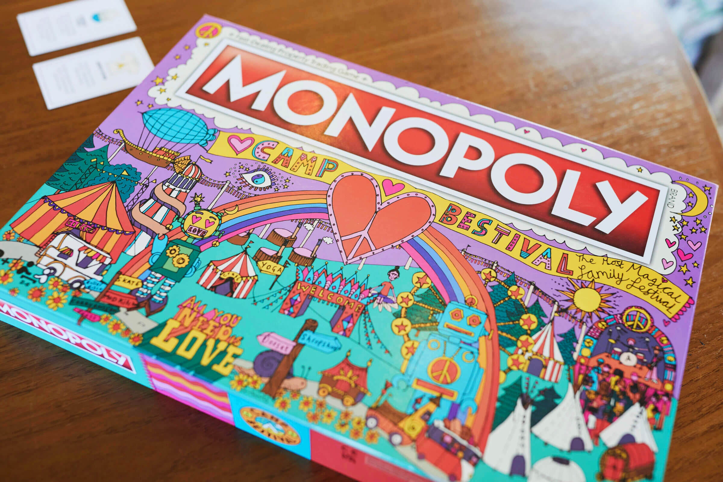 A colorful Monopoly board game box with “CAMP BESTIVAL” theme, featuring vibrant festival illustrations, tents, a hot air balloon, a rainbow, and carnival attractions on a wooden table.