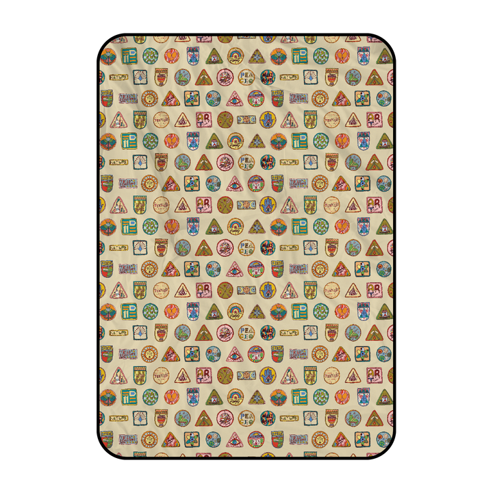The Camp Bestival Picnic Blanket by 2025 is a beige, rectangular blanket with a black border, decorated with colorful vintage-style travel badges and patches in assorted shapes like circles, triangles, and shields.